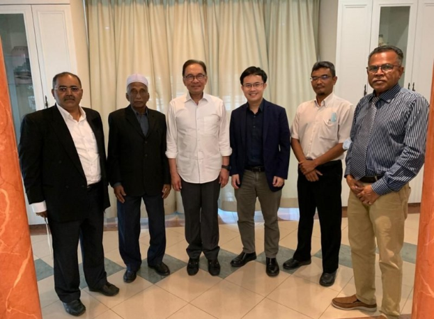 Eine Gruppe von Männern in einem formellen Raum mit zwei Säulen, Vorhängen und Schränken im Hintergrund, die in eine Diskussion vertieft sind.