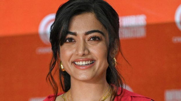 Eine Frau in einem leuchtend roten Kleid mit einem eleganten Dutt und dezentem Make-up steht lächelnd da, trägt schwarze High Heels und hat einen hellgelben Banner im Hintergrund mit der Aufschrift "Sonam Kapoors beste Schönheits Looks vom Cannes Film Festival".