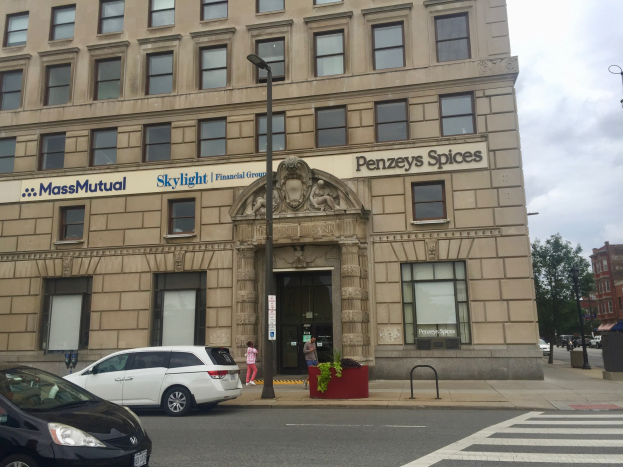 Großes Gebäude mit parkenden Autos davor, Straßenlaternen, Fahrzeugen, Fußgängern, Bäumen und Wolken, identifiziert als Hauptsitz der Massachusetts Mutual Financial Group.