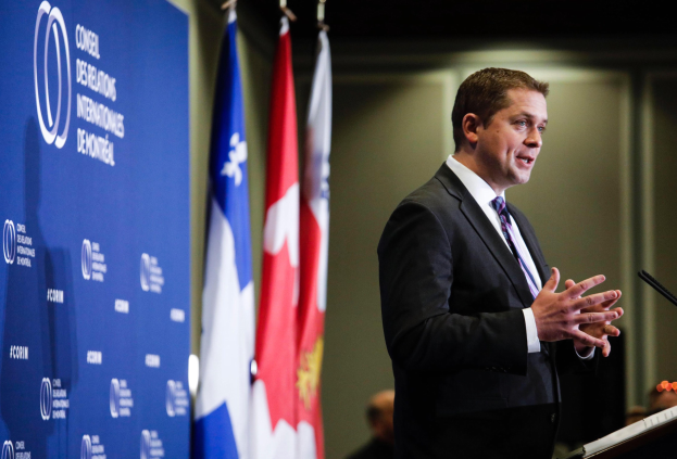 Kanadischer Premierminister an einem Rednerpult stehend, umgeben von Flaggen und einem Banner, mit Menschen und einer Wand im Hintergrund.