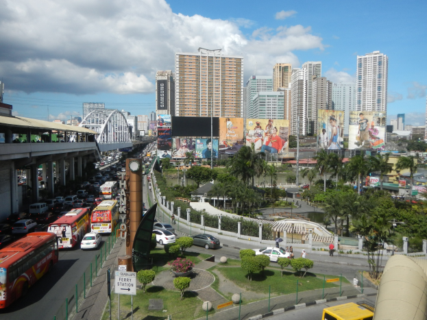 Eine belebte Stadtstraße mit Bussen, Autos und anderen Fahrzeugen, gesäumt von Bäumen, Pflanzen und Gebäuden, mit einer Brücke und einem bewölkten Himmel im Hintergrund.