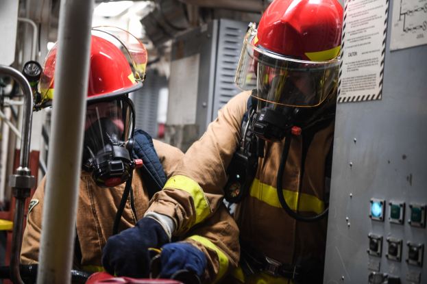 Zwei Feuerwehrleute in Schutzausrüstung arbeiten an einem Feuerwehrauto, mit einer Tafel und Metallstangen im Hintergrund.