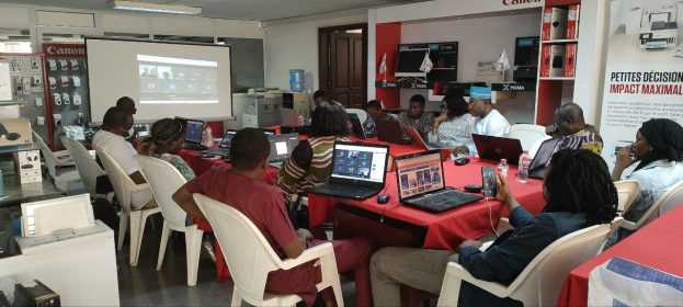 Gruppe von Menschen um einen Tisch mit Laptops und anderen Gegenständen während eines digitalen Marketing-Workshops, mit einem Banner, einem Display, einer Tür und Deckenleuchten im Hintergrund.