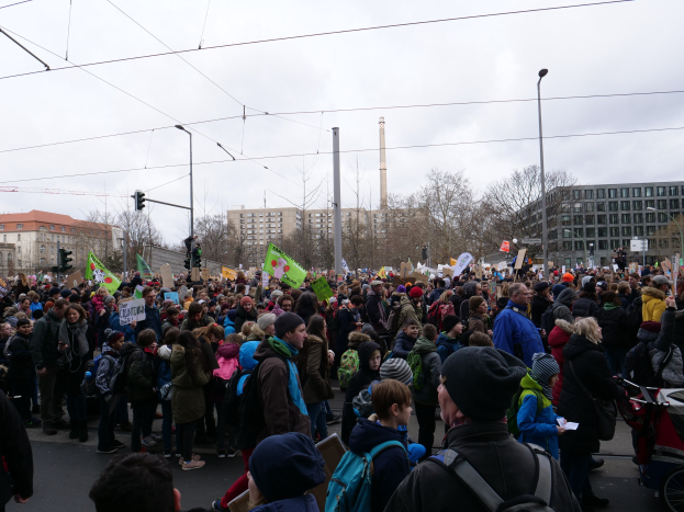Eine große Menge marschiert eine Straße entlang und trägt Schilder und Banner, einige tragen Mützen und Taschen oder schieben Kinderwagen, mit Laternenmasten, Verkehrsampeln, Bäumen, Gebäuden und einem fernen Turm unter einem klaren Himmel.