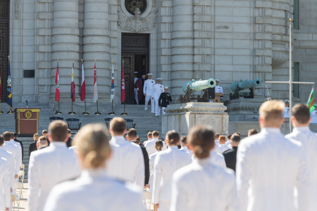 Gruppe von Menschen in weißen Marineuniformen vor einem Gebäude mit Säulen und Treppe, mit Fahnen, einem Podium, einem Mikrofon und Kanonen im Hintergrund.