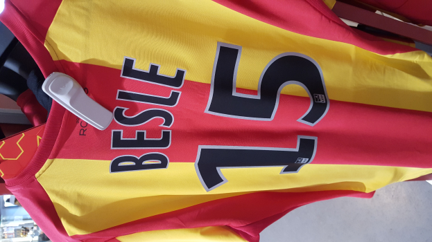 Roter und gelber Fußballtrikot mit aufgedrucktem Text und Zahlen, das an einer Ladengestell hängend mit anderen Gegenständen und dem Boden im Hintergrund.