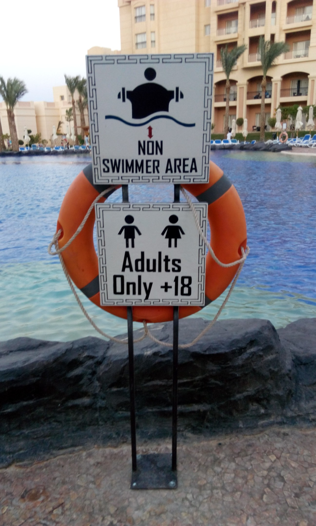 Ein Schwimmbad mit einem "Schwimmerbereich nur für Erwachsene"-Schild im Vordergrund, umgeben von Bäumen, Gebäuden und einem klaren blauen Himmel, mit einem Rettungsring in der Mitte des Pools.