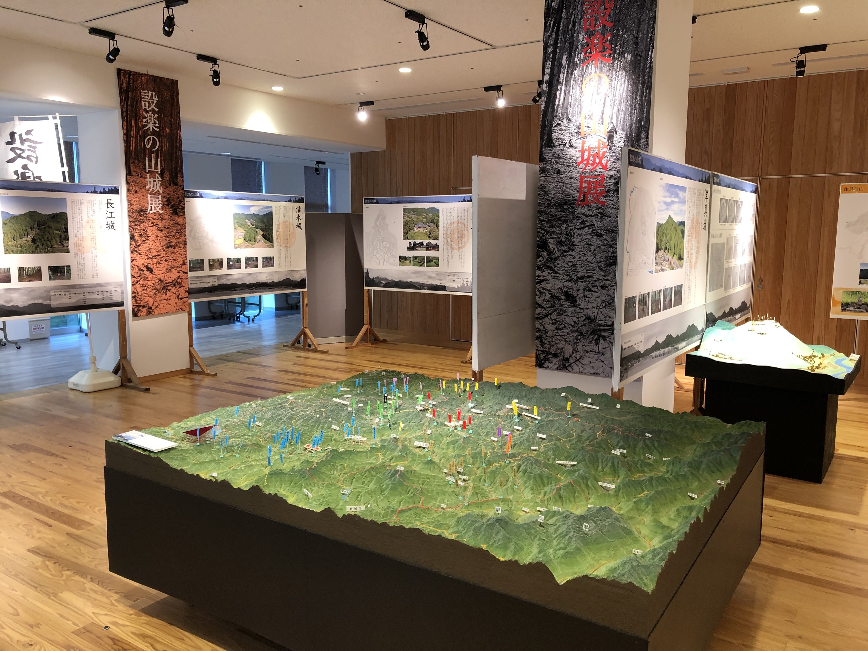 Ausstellungsraum mit verschiedenen historischen Karten Japans, einschließlich montierter Anzeigen, Bannern und einem zentralen Tisch mit einer Karte darunter, unter Deckenleuchten an einer hölzernen Wand.
