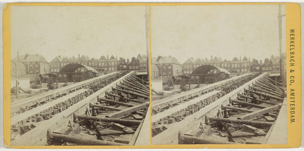 Altes Schwarz-Weiß-Foto einer Bahnschiene mit Gebäuden im Hintergrund, Text "Merkelbach & Co, Co. Amsterdam" auf der rechten Seite und verstreute hölzerne Gegenstände im Vordergrund.