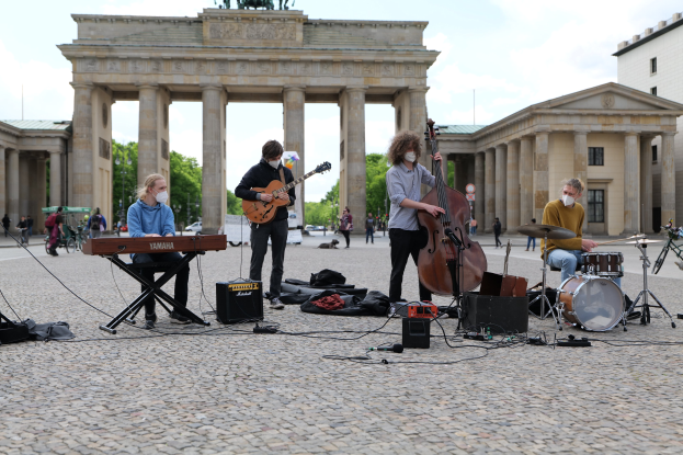 Eine Gruppe von Menschen, die Musik im Freien spielen, mit einem Gitarristen im Sitzen, einem weiteren Musiker und einer maskierten Person, vor dem Hintergrund des Brandenburger Tors in Berlin.