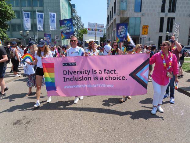 Eine Gruppe von Menschen marschiert auf einer Straße, hält ein Banner mit der Aufschrift "Diversity is a Fact, Inclusion is a Choice" in den Händen, einige tragen Mützen und Schutzbrillen, andere zusätzliche Banner, vor einem Hintergrund aus Gebäuden, Bäumen, Laternenmasten und einem klaren blauen Himmel.
