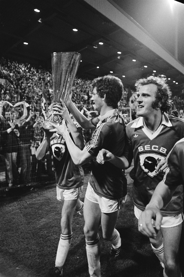 Schwarzes Bild von Fußballspielern, die einen Pokal auf einem Feld halten und feiern, mit Zuschauern hinter einem Zaun und Stadionbeleuchtung von oben.