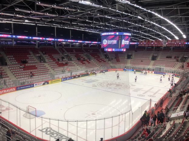 Ein Eishockeyspiel in einer großen Arena mit Zuschauern, eine von einem Zaun umgebene Eisfläche, ein Display, das "St. Louis Blues vs Detroit Red Wings" anzeigt, Metallrahmen, ein Dach mit Deckenleuchten und eine Menge.
