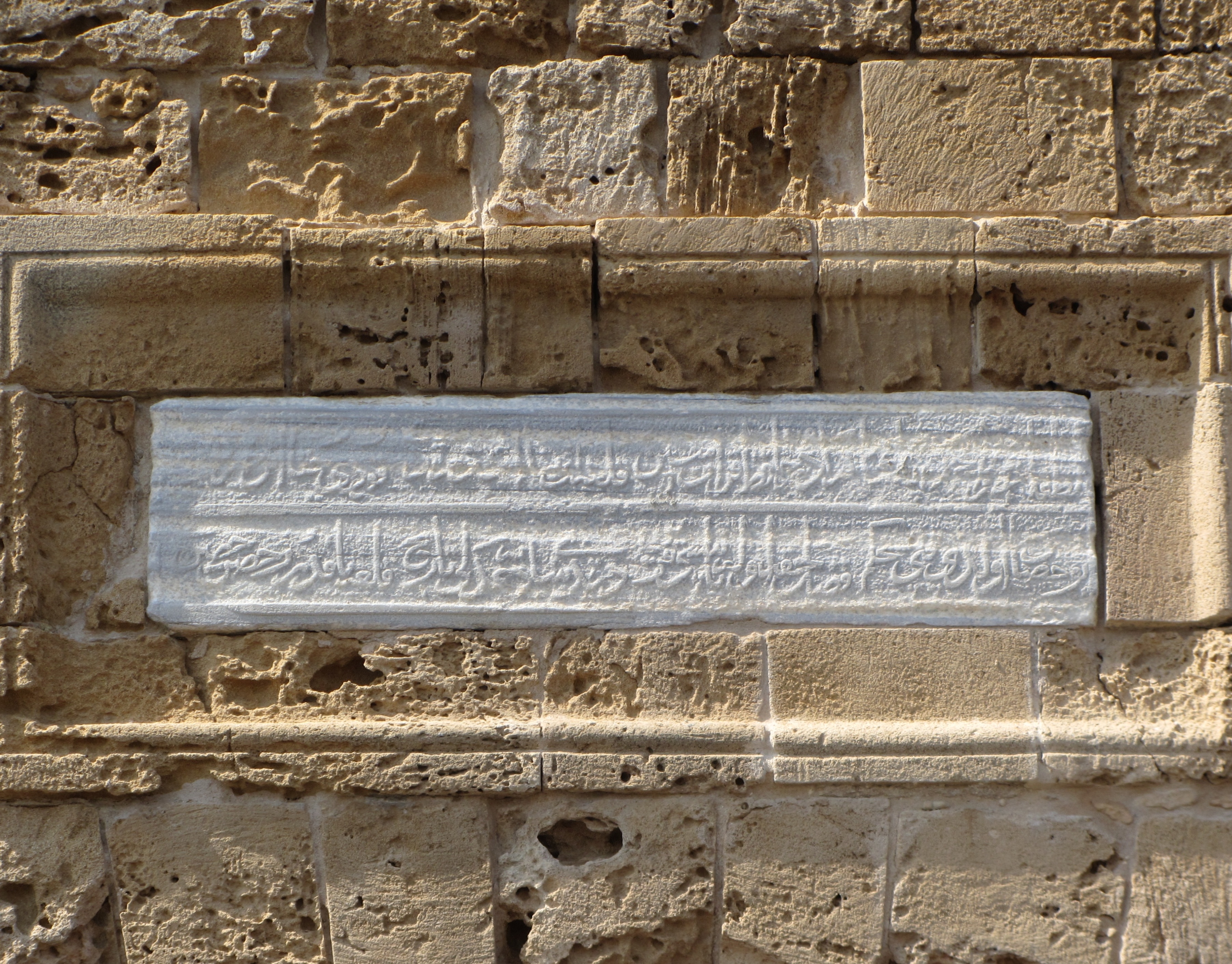 Ein Steinplakette an einem Gebäude mit arabischer Schrift und kunstvollen Verzierungen, die wahrscheinlich eine Szene aus dem islamischen Glauben darstellen, umgeben von einem dekorativen Rahmen.