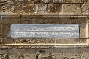 Ein Steinplakette an einem Gebäude mit arabischer Schrift und kunstvollen Verzierungen, die wahrscheinlich eine Szene aus dem islamischen Glauben darstellen, umgeben von einem dekorativen Rahmen.