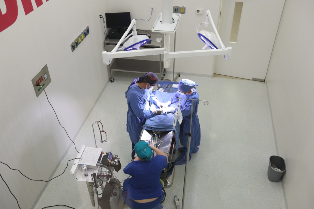 Eine Gruppe von Chirurgen führt einen Eingriff in einem Operationssaal durch, mit einem Patienten auf einem Operationstisch umgeben von medizinischer Ausrüstung, Text an der linken Wand, einem Objekt auf dem rechten Boden und verschiedenen medizinischen Geräten im Hintergrund, einschließlich eines Monitors.