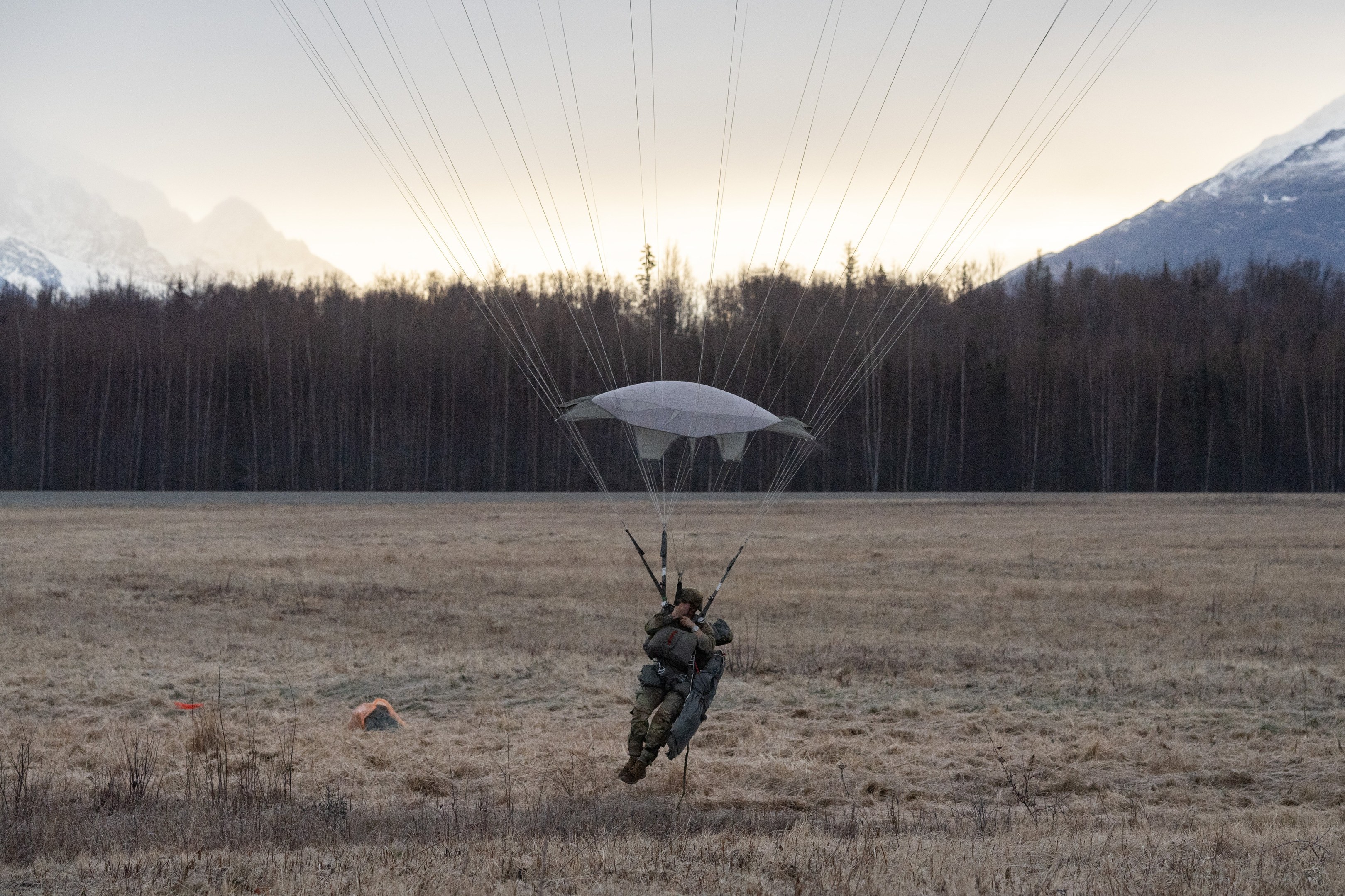 Eine Person beim Paragliding über einem Feld mit schneebedeckten Bergen im Hintergrund, die einen Helm trägt und von grünem Gras und Bäumen umgeben ist.