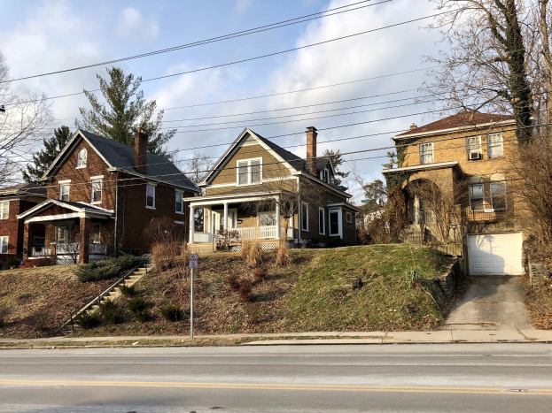 Eine Reihe von Häusern mit Fenstern, Türen, Säulen, Geländern, Stufen, Pflanzen und Gras, die entlang einer Straße mit Bäumen und einem Wegweiser in der Vordergrund aufgereiht sind, unter einem bewölkten Himmel.