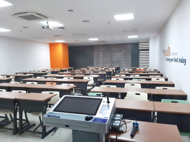 Ein Klassenzimmer mit Tischen und Stühlen in Reihen, einem deckengehängten Projektor und Gegenständen auf den Tischen sowie Text an der rechten Wand, der auf eine Privatschule in Singapur hinweist.