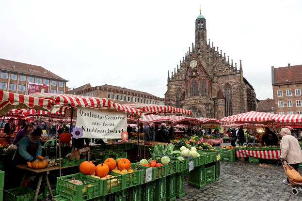 Ein belebter Markt in Nürnberg, Deutschland, mit farbenfrohen Obst und Gemüse auf dem Marktplatz, Menschen mit Taschen und um den Markt aufgestellte Zelte, mit Gebäuden und einem Uhrturm im Hintergrund unter einem sichtbaren Himmel.