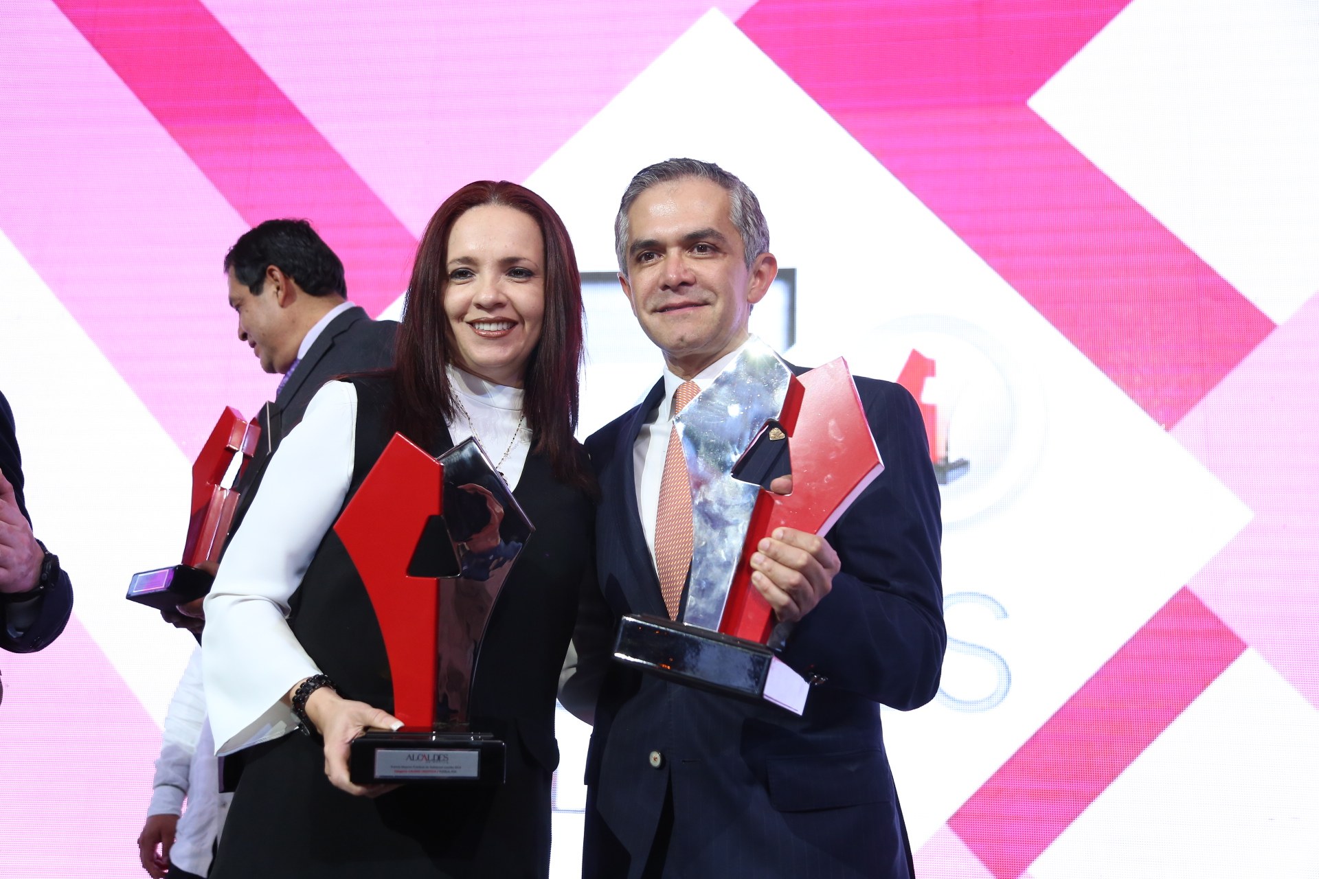 Zwei Personen auf einer Bühne, die strahlen und Trophäen halten, mit einem Bildschirm im Hintergrund.