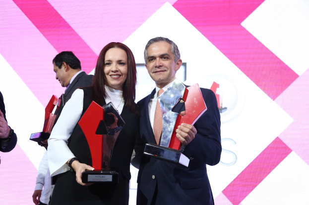 Zwei Personen auf einer Bühne, die strahlen und Trophäen halten, mit einem Bildschirm im Hintergrund.