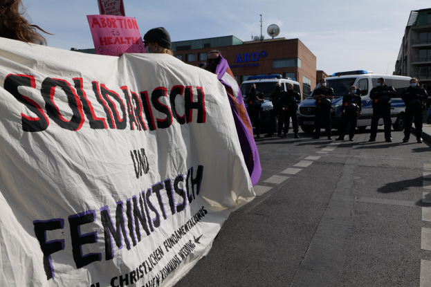 Eine Gruppe von Menschen marschiert auf einer Straße, trägt ein Banner mit der Aufschrift "Solidarität und Feminismus", mit parkenden Fahrzeugen und Gebäuden im Hintergrund unter einem klaren blauen Himmel.