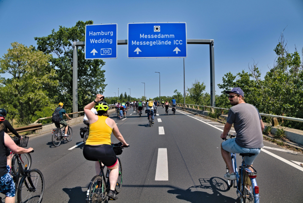 Eine Gruppe von Radfahrern mit Helmen fährt auf einer Straße mit einer Begrenzung auf einer Seite und Bäumen auf der anderen unter einem klaren blauen Himmel mit Laternen im Hintergrund; ein Schild oben zeigt eine Radtour in Hamburg an.