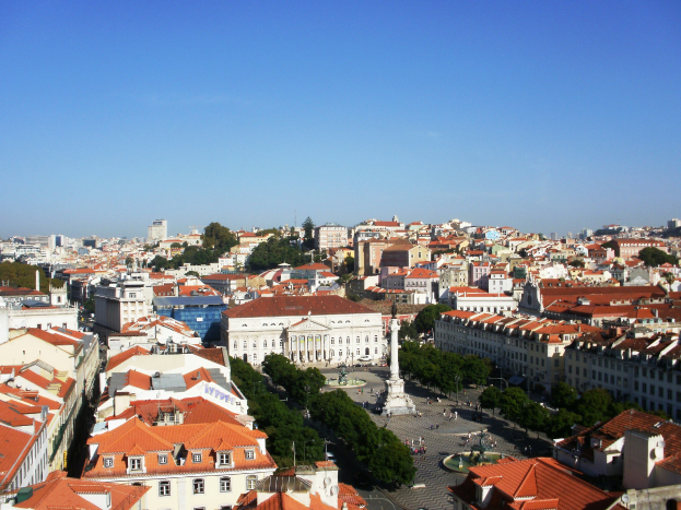 Blick auf Lissabon von einem Hügel aus, der Gebäude mit Fenstern, Bäume, eine Statue auf einem Sockel, Menschen auf der Straße und den Himmel im Hintergrund zeigt.