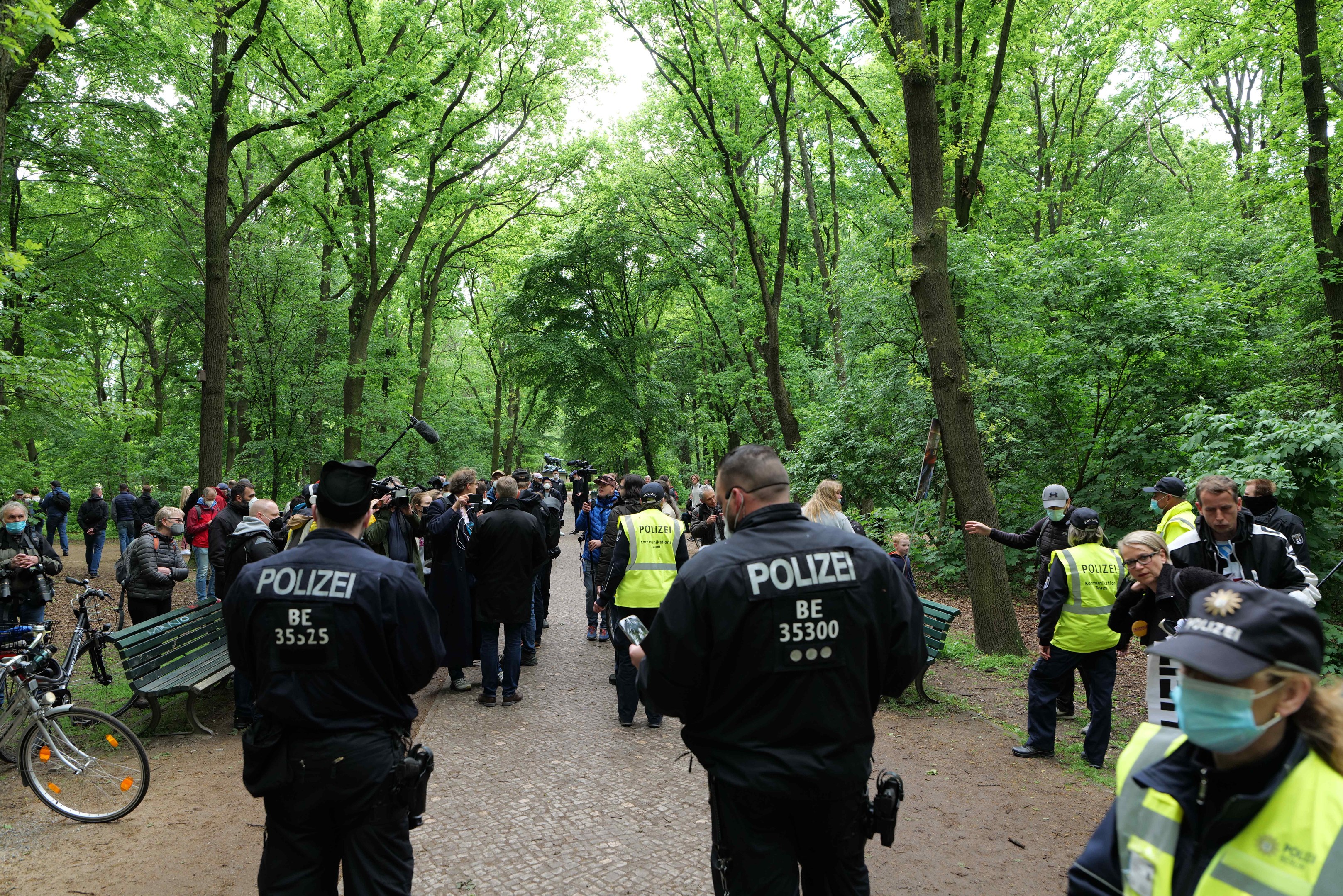 Polizisten vor einer Menge während einer Anti-Terror-Demonstration in Berlin, mit Fahrrädern und einer Bank im Vordergrund und Bäumen im Hintergrund.