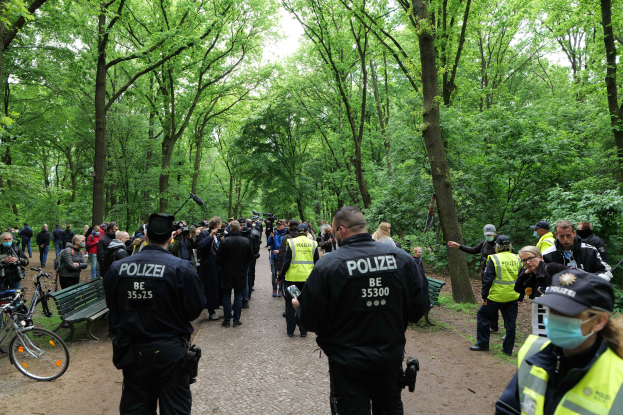 Polizisten vor einer Menge während einer Anti-Terror-Demonstration in Berlin, mit Fahrrädern und einer Bank im Vordergrund und Bäumen im Hintergrund.