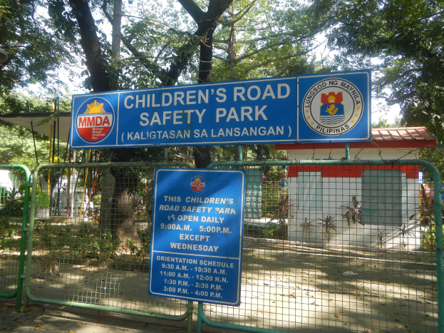 Ein von einem Metallzaun umgebener Kinderspielplatz für Verkehrssicherheit mit zwei blauen Informationsschildern, einem kleinen Haus mit Dach, Topfpflanzen, einer Baumgruppe und einem bewölktem Himmel.