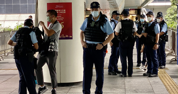 Eine Gruppe von Polizisten in Uniform und mit Mund-Nasen-Schutz steht vor einem Gebäude, einige halten Kameras, mit einem Banner, Geländern, Pflanzen, Bäumen und Deckenleuchten im Hintergrund.