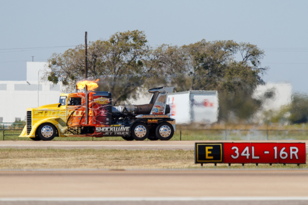 Ein Lkw mit Flammen fährt eine Straße entlang mit Gras, einem Zaun, Bäumen, Polen, Drähten, einem Gebäude und einem Himmel im Hintergrund.