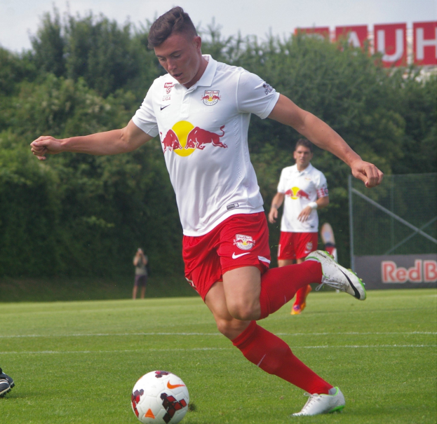 Ein professioneller Fußballspieler schießt den Ball auf einem Rasenfeld mit Bäumen und einem klaren blauen Himmel im Hintergrund, wobei "RB Leipzig" auf einem Schild zu sehen ist.