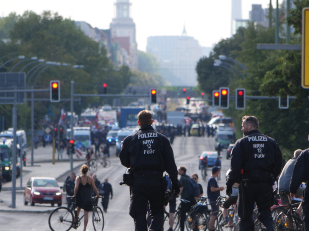 Polizeibeamte mit Fahrrädern auf einer von Bäumen gesäumten Straße mit Gebäuden und einem klaren blauen Himmel im Hintergrund.