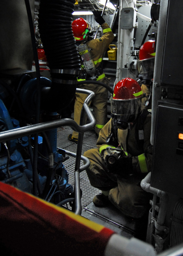 Feuerwehrleute in Helmen und Schutzausrüstung an einem Feuerlöschwagen mit Rohren und einer Lampe im Hintergrund.