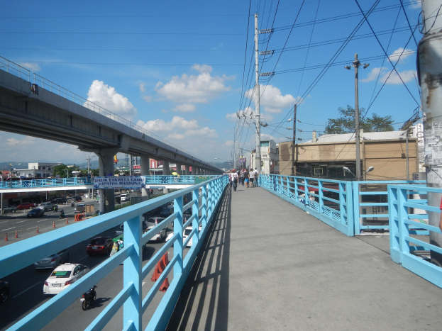 Eine Fußgängerbrücke mit Menschen darauf, darunter Fahrzeuge und im Hintergrund Gebäude, Bäume und Himmel; eine Person steht rechts auf der Brücke.