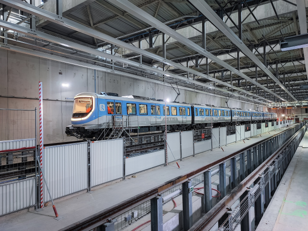 Ein blau-weißer Zug fährt durch eine große Bahnhofshalle mit einer Plattform auf der rechten Seite und einer Wand im Hintergrund, unter einer Decke mit Stangen und Lampen.