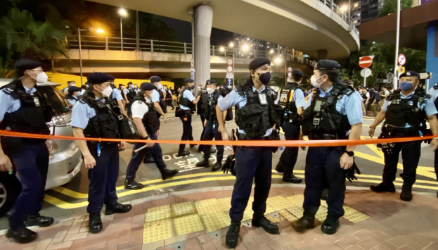 Polizeibeamte in Uniform und mit Masken vor einer Menge, mit Fahrzeugen, Gebäuden, Bäumen und einer Brücke im Hintergrund, wahrscheinlich nach einer Hongkonger Demonstration, bei der ein Mann erschossen wurde.