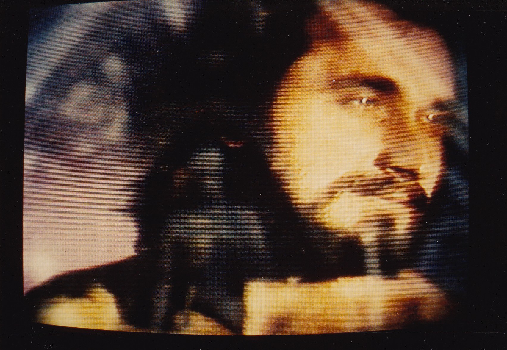 Ein Mann mit dunklem Bart und Schnurrbart, langen dunklen Haaren und einem ernsten Gesichtsausdruck in einem weißen Hemd ist auf einem Fernseher mit dunklem Hintergrund zu sehen.