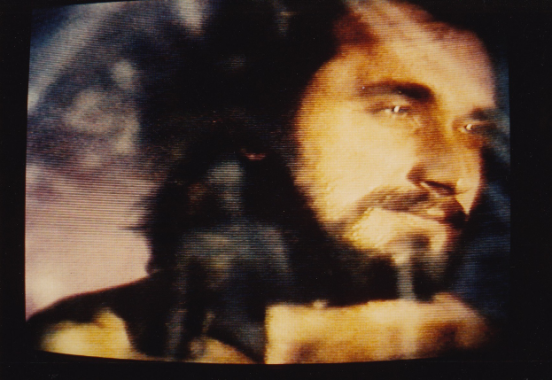 Ein Mann mit dunklem Bart und Schnurrbart, langen dunklen Haaren und einem ernsten Gesichtsausdruck in einem weißen Hemd ist auf einem Fernseher mit dunklem Hintergrund zu sehen.