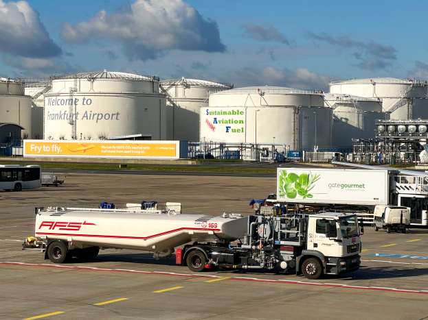 Ein großer weißer und grüner Tanklastzug parkt vor einem Gebäude am Frankfurter Flughafen, umgeben von anderen Fahrzeugen, großen weißen Tanks und einem gelben Schild im Hintergrund bei klarem blauem Himmel.