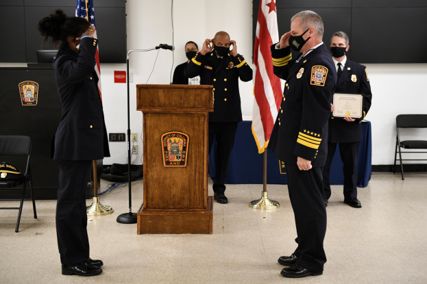 Gruppe uniformierter Personen in Masken vor einem Podium mit Mikrofon, umgeben von Fahnen und Möbeln, während einer Vereidigung eines neuen Feuerwehrchefs, wobei eine Person ein Zertifikat hält.