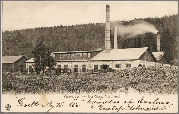 Altes Schwarz-Weiß-Foto einer Fabrik, die Rauch aus ihren Schornsteinen ausstößt, mit Häusern, Bäumen, Pflanzen und einem Zaun drumherum und Text unten.