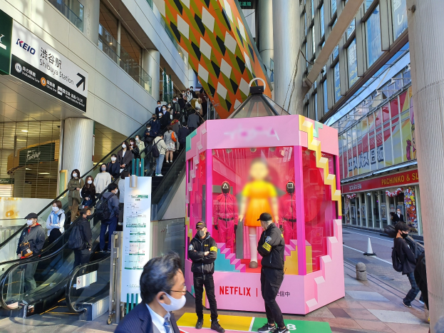 Eine Gruppe von Menschen steht vor einem Gebäude mit Glaswänden, Säulen und Texttafeln, einige tragen Masken, während daneben eine Rolltreppe mit Menschen hinauffährt; in der Mitte eine pinkfarbene Box mit Schaufensterpuppen, die den Text 'Netflix's new Netflix booth in Tokyo' anzeigt.