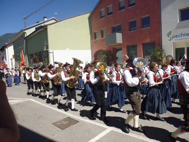 Eine Gruppe von Menschen in traditioneller bayrischer Tracht, die auf der Straße Musikinstrumente spielen, während sie durch eine Straße mit Gebäuden gehen, einige halten Fahnen, mit einem Hügel und einem klaren blauen Himmel im Hintergrund.