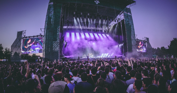 Eine große Menschenmenge bei einem Musikfestival mit einer Bühne, auf der Musiker spielen, beleuchtet von Lichtern und Bildschirmen, vor einer Kulisse aus einem Gebäude, Bäumen und einem klaren blauen Himmel.