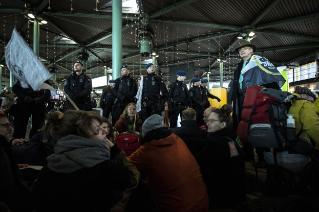 Eine Gruppe von Menschen steht vor einer protestierenden Menge in einem Bahnhof, mit Säulen und Deckenleuchten im Hintergrund.