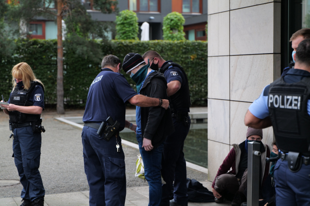 Eine Gruppe von Polizisten vor einem Gebäude stehend, mit einer Frau mit Buch und Stift, sowie mehreren Personen in der Nähe sitzend und stehend, vor einem Hintergrund aus Grünflächen und städtischen Strukturen.